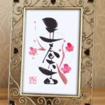 筆文字『立春大吉』作品。縦書きで左右対称の四文字に梅の花を優しくあしらった春の厄除け開運アート。玄関飾りにおすすめ。